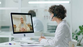Female doctor in headset and white coat sitting in clinic and having discussion with eldelry woman via online video call on computer during online consultation