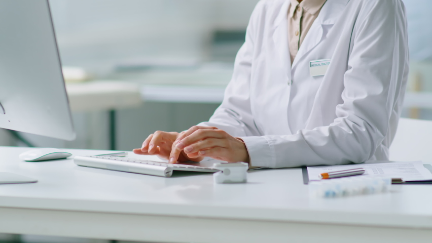 Tilt up shot of female doctor in white coat typing on computer while working at desk in medical office