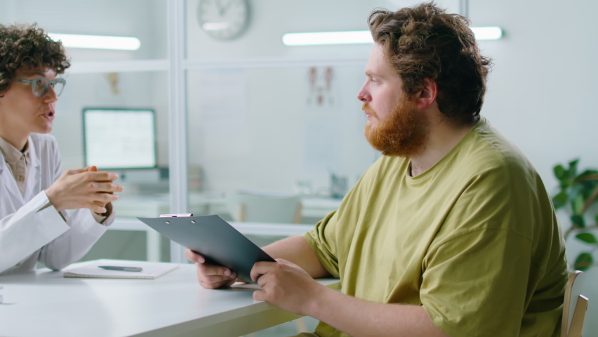 Young overweight man giving medical history to female doctor and getting consultation about weight loss in clinic Royalty-Free Stock Footage #1092211319