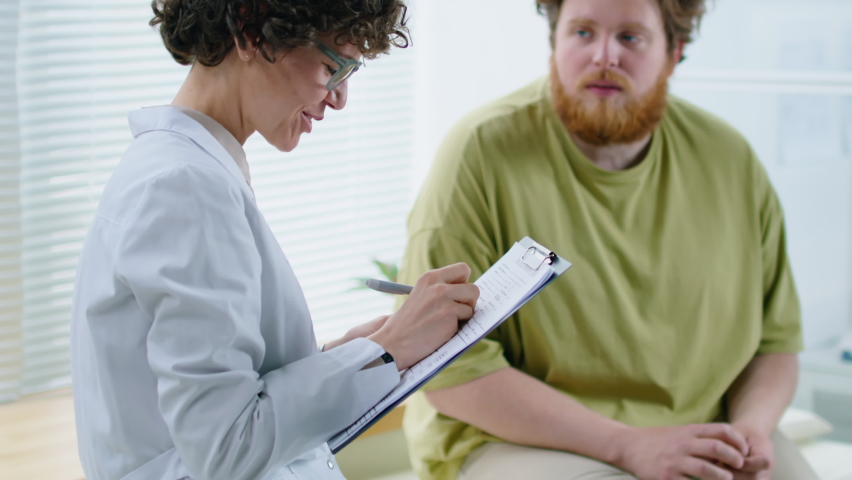 Female dietician speaking with overweight male patient and taking notes during consultation in medical office | Shutterstock HD Video #1092211709