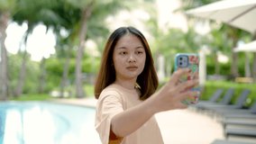 4K, A close-up shot of a young Asian tourist taking a selfie with her cell phone happily at the poolside of her hotel on a weekend.