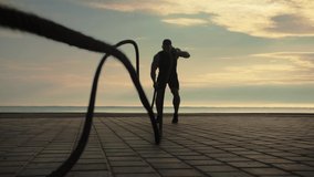 Bodybuilder training outdoors doing an exercise with battle ropes against the backdrop of sunrise. Training lifestyle.Slow Motion