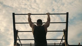 Athletic man is training outdoor, doing pull-ups on his back muscles. Against the background of the sky in the clouds.