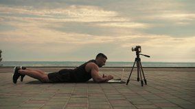Аthletic man looks at a laptop lying on a mat, writing an online workout program against a backdrop of sunrise and the sea.