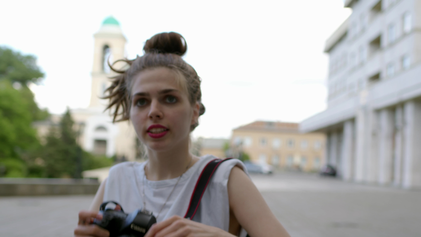 professional photographer in white T-shirt and with bun on her head holds black camera in hands and takes picture. woman take photos and adjusts camera, look into viewfinder