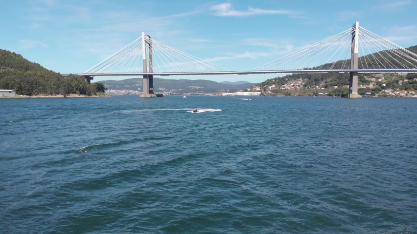 jet ski makes its way past the Puente de Rande, Rande Bridge located in the city of Vigo, Galicia, Spain. The world-famous Ría de Vigo known for mussels and beautiful summer weather