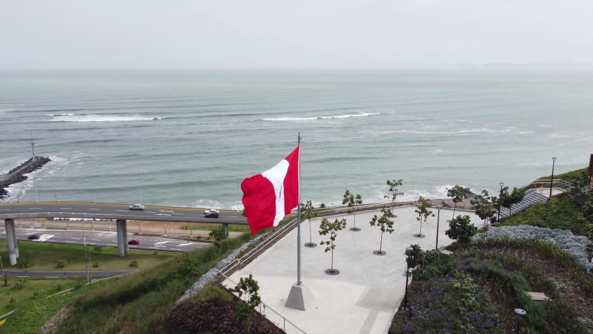 Drone video of a park on edge of a cliff in Lima, Peru in Miraflores district. Drone flies forward over a big peruvian flag. Down below are coastal freeway streets and then the beach and ocean. Royalty-Free Stock Footage #1092226547