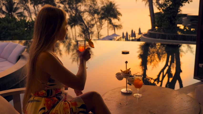 Young woman drinks cocktail sitting in luxury outdoors restaurant at hotel or resort at evening sunset. Girl in evening dress holds mocktail. Summer holidays or vacations, travel, tourism concept. | Shutterstock HD Video #1092228151