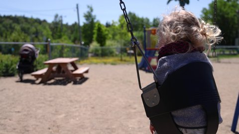 An excited toddler is seen from behind, sitting in a toddler swing and raising hands in air swaying back and forth. Blurry playground in background.