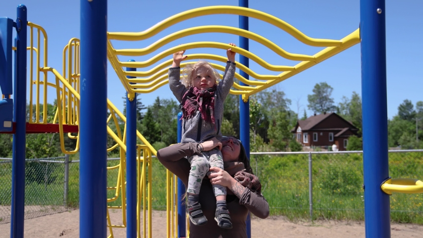 Slow motion video from the front as happy mother lifts two year old son up, so he can reach the tall monkey bars of a climbing frame on a park. Royalty-Free Stock Footage #1092229575