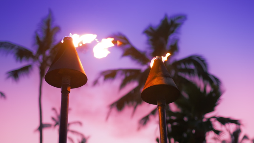 Tiki torches on Hawaii in slow motion. Torches with fire and flames burning in dramatic Hawaii sunset sky by palm trees. Beautiful pink purple sunset sky with torches on Hawaiian Waikiki beach, Oahu