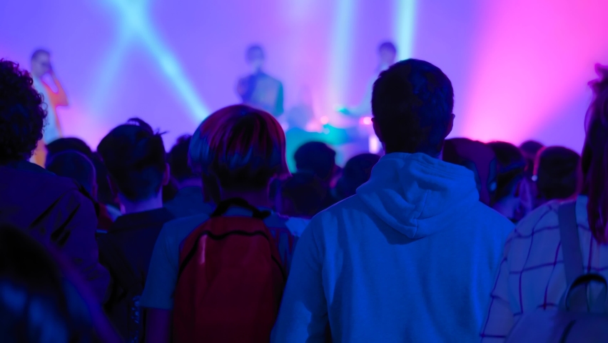 Slow motion: teenagers partying, cheering, raising hands up and jumping at rock concert in front of stage of nightclub. Bright colorful stage lighting. Nightlife and entertainment concept