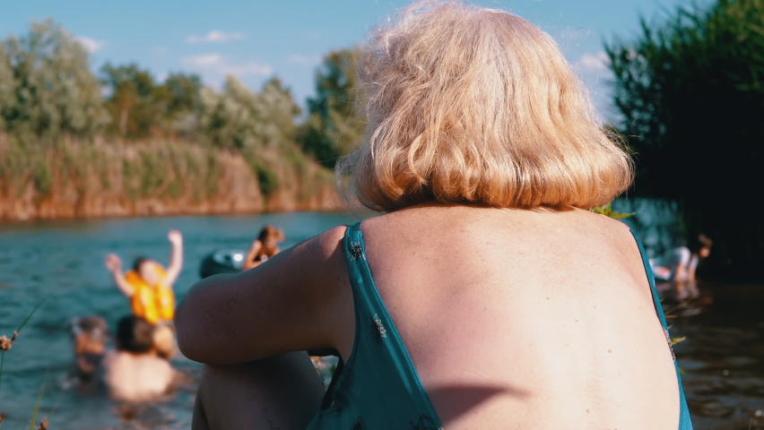 Elderly Woman on Beach in a Bathing Looks at the Bathing Children in the River. Back view. Female enjoys nature on bank of the river, blue sky, lake, reeds, looks at the floating children. Weekend.