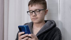 Smart Boy in Glasses Watching a Video in a Mobile App on a Smartphone. A serious teenager in a bathrobe plays video games and writes messages in room. Online learning. Internet addiction. Lifestyles.