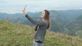 Young woman stands on mountain, video communication on smartphone, waves hand, says hello. Nice view mountains, clouds. Concept of internet connection, online communication, selfie, nature, goal