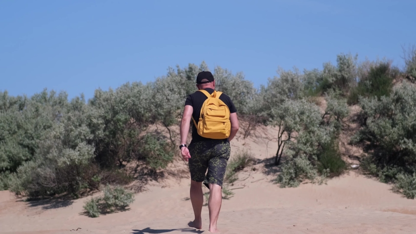 Man with backpack hiking in beautiful coastal sand dune towards beach. Barefoot male traveler walking on the sandy desert surface with vegetation, blue sky on background. Vacation, adventure concept