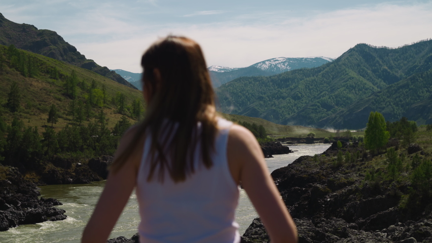 River runs across mountain valley and woman tourist on sunny day backside view. Wild environment of hillside in windy weather. Wanderlust lifestyle