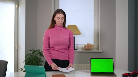 Portrait of unsatisfied young Caucasian woman looking at camera talking with green screen laptop on table with web page mockup. Dissatisfied freelancer posing explaining startup failure