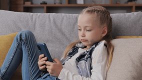 Portrait of surprised little cute girl holding smartphone lying at sofa at home. Choosing favorite music or cartoons,texting messages,browsing internet,watching video, playing games on smartphone