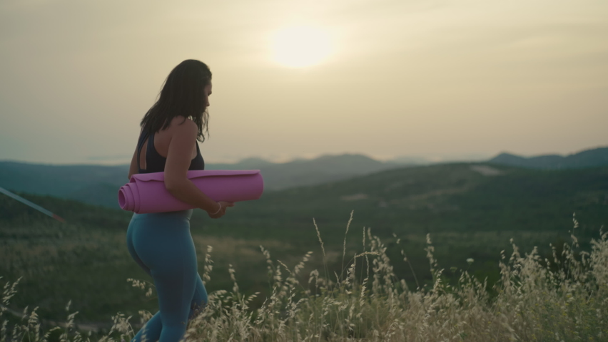The girl with a rug in her hands goes to the top of the mountain to practice yoga. Fitness and healthy lifestyle woman in the mountains at sunset.