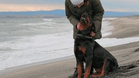 Unknown girl in warm jacket stands on sandy beach near blue stormy sea, and scratches behind ear of her faithful friend - large beautiful educated dog of Rottweiler breed, 4K UHD slow-motion video