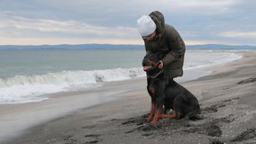 Unknown girl in warm jacket stands on sandy beach near blue stormy sea, and scratches behind ear of her faithful friend - large beautiful educated dog of Rottweiler breed. 4K UHD slow-motion video