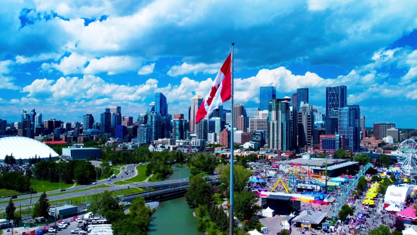 Calgary Stampede arial drone hold horizon city skyline sunny summer day largest Canada flag in the foreground MNP community sports Repsol Centre divided by river running lush green thousands of people Royalty-Free Stock Footage #1092258239