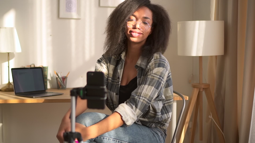 Portrait of young woman is talking in front of smartphone and sitting in home room . Close view of African American woman turns on broadcast and looks at display of phone, speaks with smile and