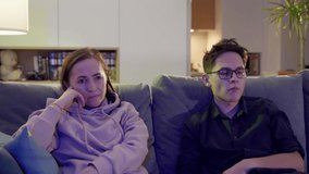 Woman with adult son watching TV sitting on the couch. A young man in glasses sits with his mother on the couch and eats popcorn.