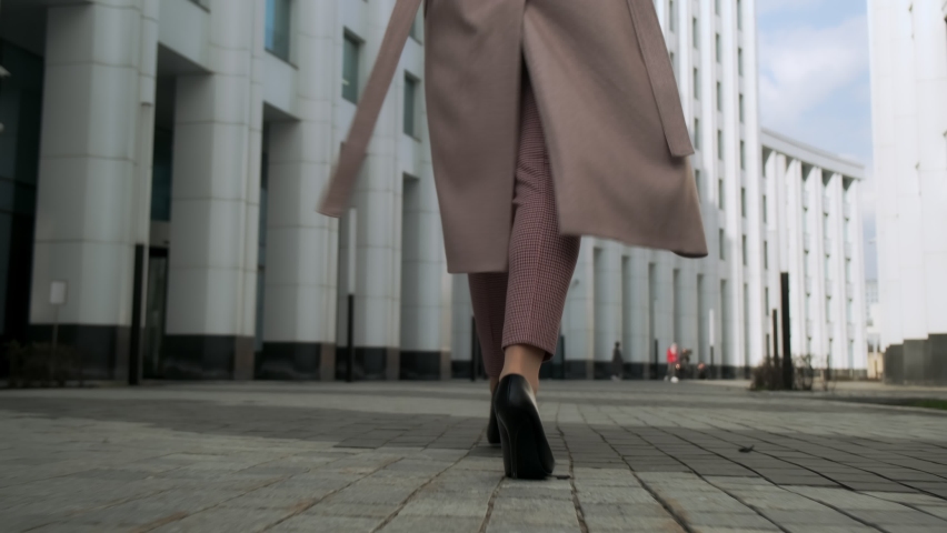 Business female legs in heels, in trousers with a developing coat, walks through the business center. Back view.