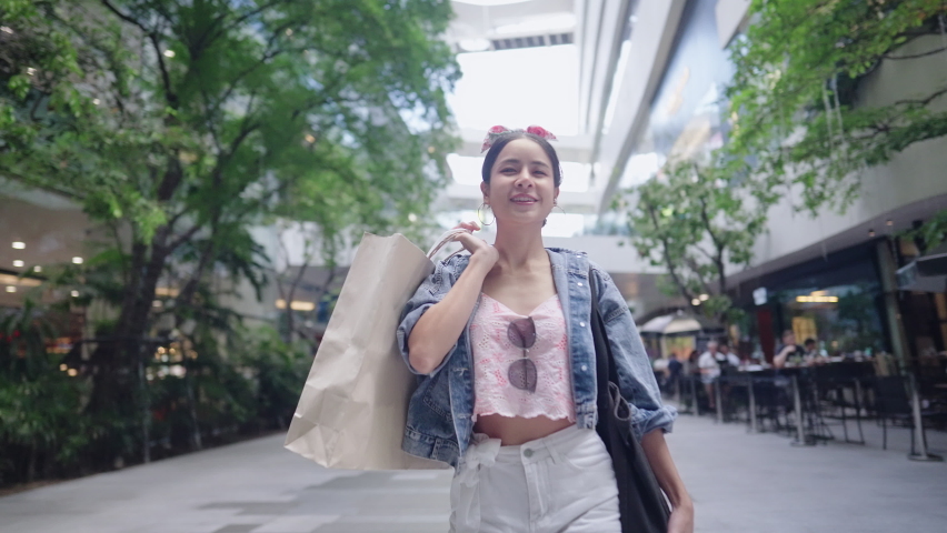 Handheld Shot on Attractive fashionable tourist woman enjoys lifestyle inside community mall with paper shopping bags in hands. Eco friendly, sustainable building with vertical garden in modern city.  Royalty-Free Stock Footage #1092268727