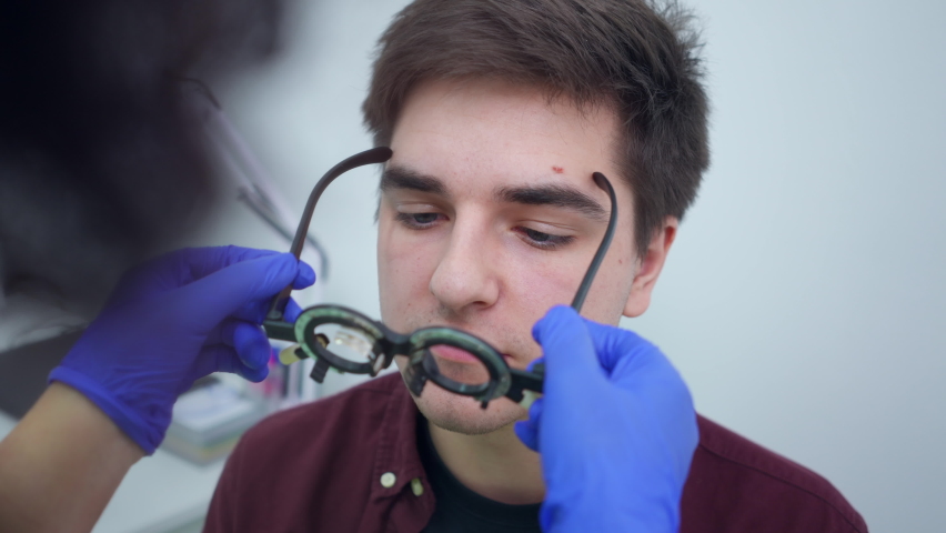 Close-up portrait of young Caucasian man with bad eyesight sitting in hospital as unrecognizable doctor putting on trial frame with lens. Headshot of patient undergoing medical examination indoors