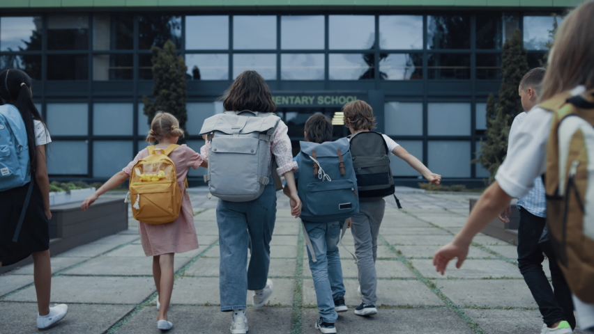 Diverse children going school with backpacks back view. Cheerful smiling teachers meet welcoming teen students at open door. Excited elementary age pupils entering modern building. Study concept. Royalty-Free Stock Footage #1092280759