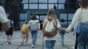 Diverse children going school with backpacks back view. Cheerful smiling teachers meet welcoming teen students at open door. Excited elementary age pupils entering modern building. Study concept. - Powered by Shutterstock - Get 15% off with code: PIKWIZARD15
