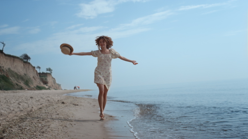 Happy cheerful girl jumping on water sandy beach summer day. Attractive curly woman running on ocean waves waving straw hat. Smiling cheerful lady have fun on seashore enjoy beautiful nature.
