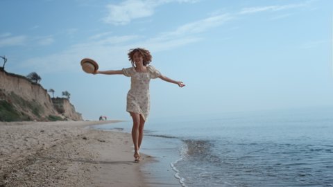 Happy cheerful girl jumping on water sandy beach summer day. Attractive curly woman running on ocean waves waving straw hat. Smiling cheerful lady have fun on seashore enjoy beautiful nature. Adlı Stok Video