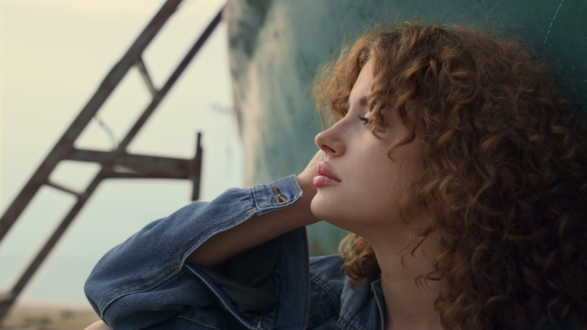 Dreamy gorgeous woman sitting beach gloomy evening leaning on old abandoned boat close up. Attractive curly girl looking camera seductively holding hand on face. Portrait of relaxed young model. Royalty-Free Stock Footage #1092280863