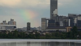 A bright rainbow is visible in the sky over a large modern city. Skyscrapers are reflected in the waters of a large reservoir.