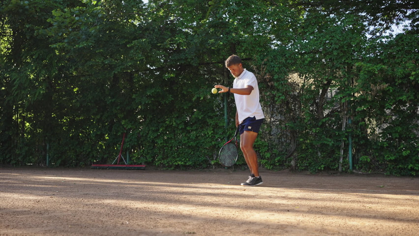 Young man serves hitting ball jumping on sand court surrounded by green hedge. Male tennis player wearing white polo t-shirt kicks ball with racquet closeup