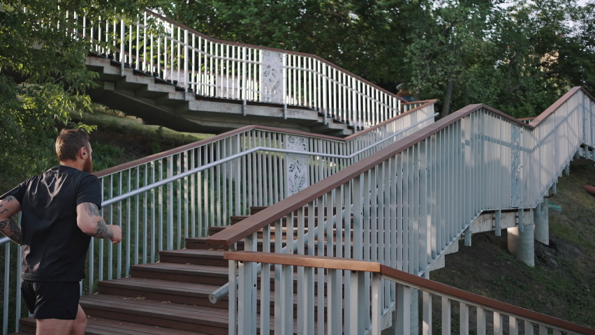 Sportive man in black sportswear runs on wooden stairs of footbridge near city park with lush trees. Athlete enjoys sports activity in summer slow motion