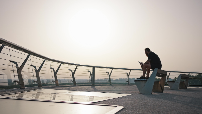 Man uses phone sitting on bench back at city viewpoint against bright sun in early morning. Athlete rests after intensive training and drinks water