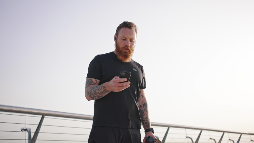 Bearded man looks in smartphone drinking water from thermos after intensive training. Mature athlete checks time standing on footbridge low angle shot
