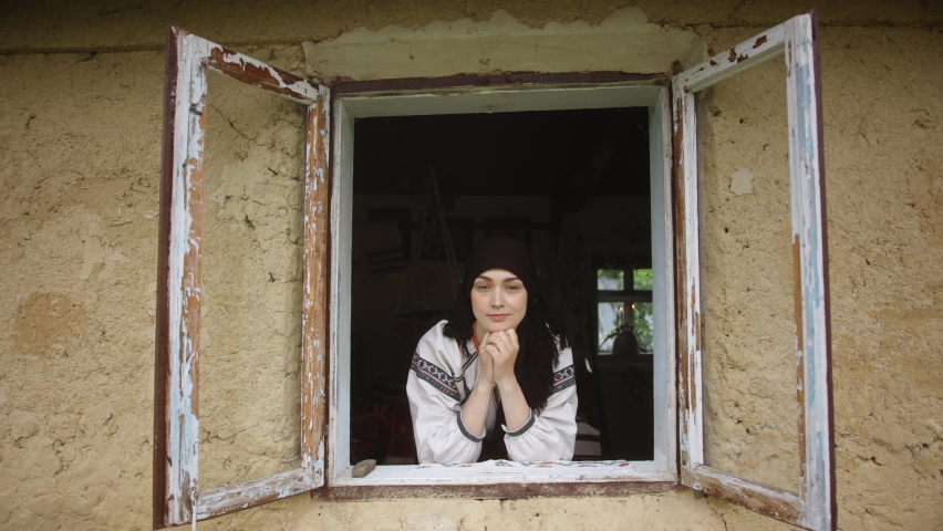 Young woman looks out of old window in ethno museum house. Young woman looks out of old window in ethno village house. Adult lady wearing Ukrainian national outfit goes smiling and sits down at table