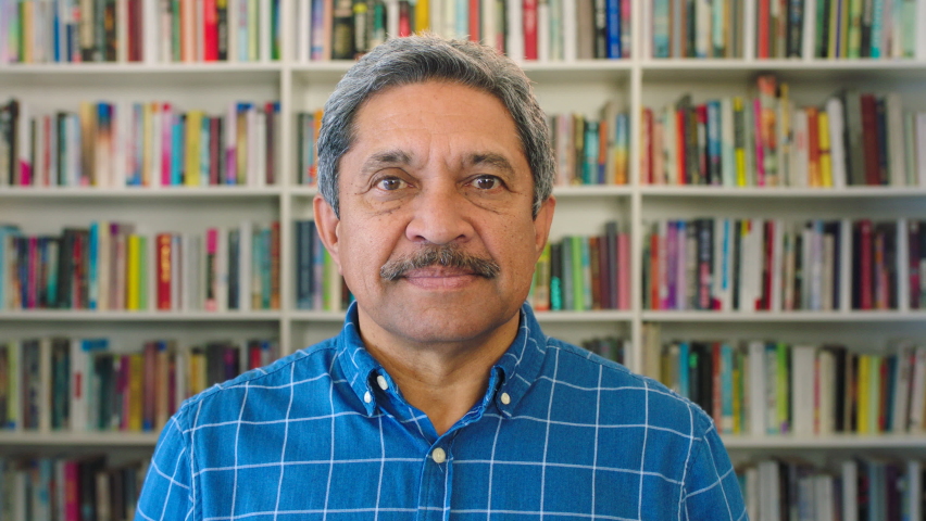 Portrait of an older male teacher or librarian looking happy, alone in a school or community library. Senior latino bookstore owner welcoming customers. Mature smiling man standing inside with books Royalty-Free Stock Footage #1092287011
