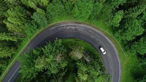 Aerial view of mountain twisted road through the forest and driving car. Mountains in summer and curved road through forest. Epic drone footage of highway in the wild mountain scenery. 