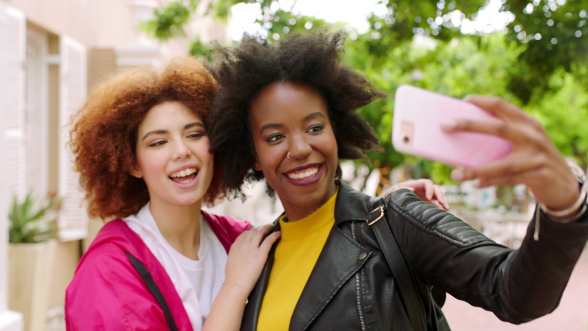 Playful female friends taking selfies with a phone outside in an urban park. Silly african girls pulling faces to post on social media. Young women smiling and laughing together making memories. Royalty-Free Stock Footage #1092296509