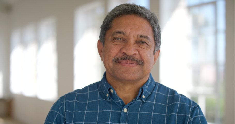 Portrait of a happy retired male looking cheerful standing inside a bright room. Face of a laughing mature latino man standing in an empty office or home. Smiling confident senior enjoying retirement | Shutterstock HD Video #1092296529