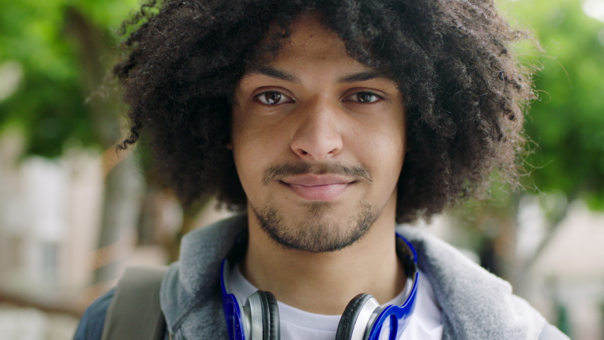 Trendy Afro American man smiling outdoors against blur background. Portrait of a confident African male with curly hair and headphones standing outside in a park or in an urban city Royalty-Free Stock Footage #1092297161