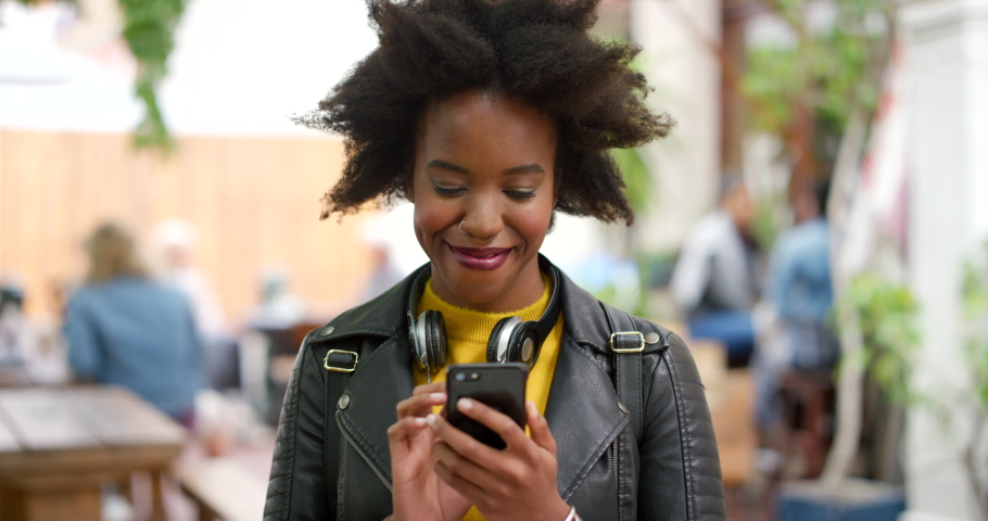 Young happy, trendy and stylish woman using a phone to send a text online in the city. One cheerful, edgy and carefree black female with an afro smiling and browsing the web while standing downtown Royalty-Free Stock Footage #1092305213