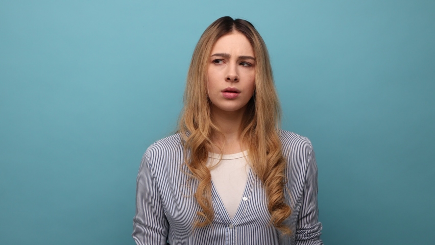 Portrait of thoughtful upset woman pondering serious issues, looking with uncertain hesitant expression, making difficult choice, wearing striped shirt. Indoor studio shot isolated on blue background. Royalty-Free Stock Footage #1092306401